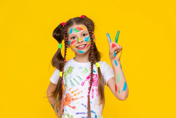 A young girl stained in multicolored paint shows two fingers and smiles broadly. Children's creativity. Yellow isolated background.