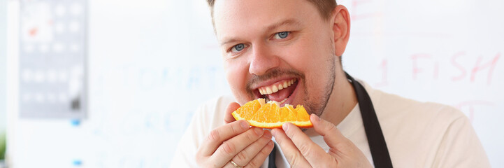 Red smiling male cook is biting orange. Fruits and healthy food concept