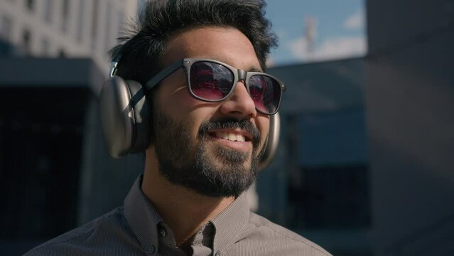 Carefree Indian Arabian businessman business man student wear sunglasses listen music sunny outdoors enjoy sound in wireless headphones near city office portrait relaxed happy guy smile in earphones 