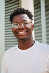 Vertical portrait of one young african american teenager smiling and looking at camera standing outdoors. Front view of happy and joyful man with eyeglasses and white perfect teeth. Lyfestyle concept