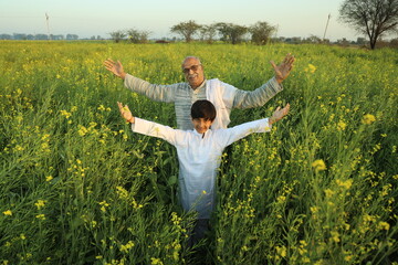 Happy Indian farmer standing in the mustard field with grand son and enjoying the benefits from the flourished rural mustard crop