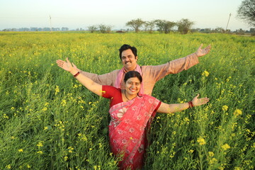 Naklejka na ściany i meble Happy Indian farmers standing in the mustard field and enjoying the benefits from the flourished rural mustard crop. husband and wife are happy to see the crops.