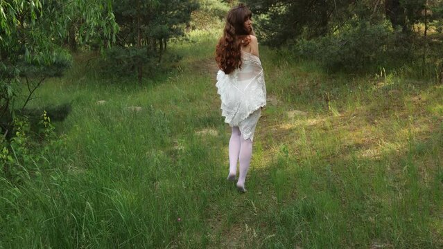 Rear view of a young woman with long hair in white lingerie walking along the road in the forest. Slow motion woman underwear