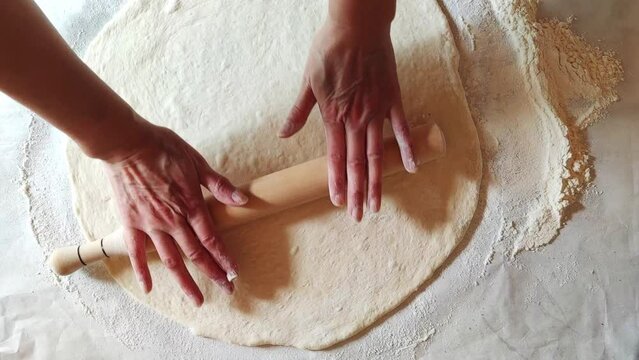 Women's hands roll out the dough with a wooden rolling pin on the kitchen table. The process of cooking at home. Top view.