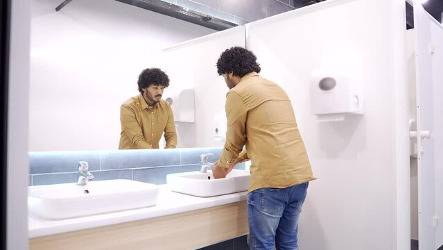 A young man washes his hands in a public toilet. Businessman in a shirt opened the tap with water in the wash basin, washes himself and looks in the mirror. Concept of personal cleanliness and hygiene