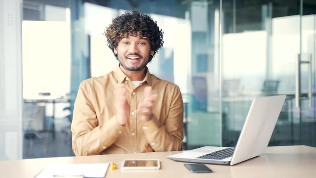 Young smiling man applauding while sitting at laptop at workplace in office and looking at camera. Positive friendly businessman clapping hands expressing positive emotions and compliment performance