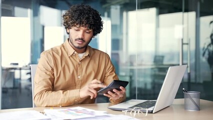 A busy young accountant calculates a project budget on a calculator, noting indicators while sitting at a desk in a modern office. Confident financier working on a financial report, doing paper work