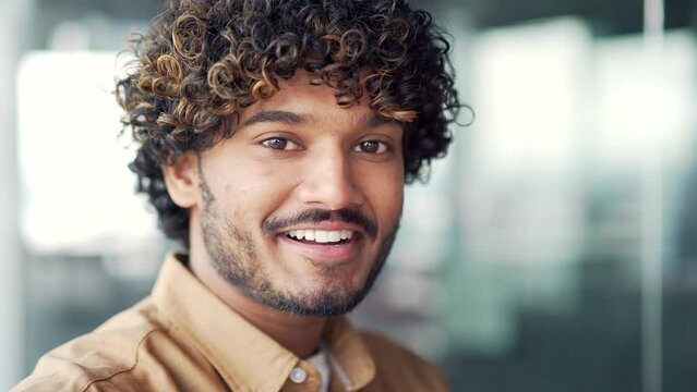 Close up portrait of young positive smiling handsome man in modern office. Happy male employee in a shirt with curly hair with a friendly look looking at the camera. Head shot of a businessman