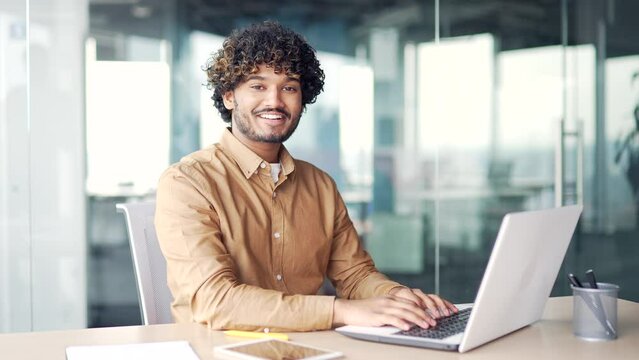 Portrait of a young successful handsome man sitting at a laptop at a workplace in a modern office. Confident smiling entrepreneur with a friendly look looking at the camera. Head shot of a employee