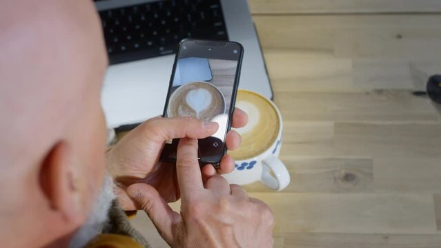 The old man's hands are holding a smartphone, the elder takes a photo of cappuccino coffee on the phone camera. Makes a blog about food or coffee shops. Senior man takes pictures on his phone. Closeup