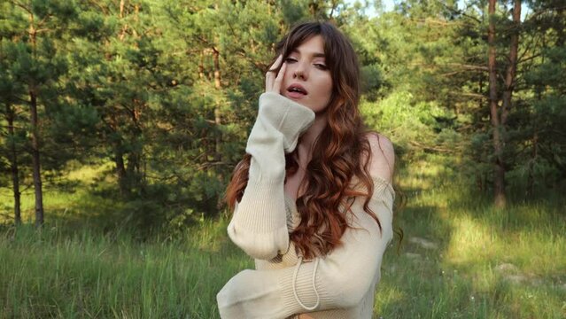 A young beautiful brunette woman with long hair in a beige tank top poses beautifully against the backdrop of a green forest. Slow motion portrait woman erotic touch face