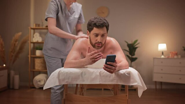 Zooming in on man with earpods having videocall. Bearded male patient actively communicating with someone while having massage in spa salon. Massagist distracting her client during his conversation.