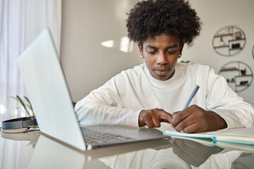 African American teenage boy student using computer watching webinar, learning english online virtual elearning class, taking english web course writing notes sitting at home table.