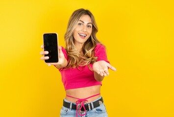 Young beautiful blonde woman wearing pink crop top over yellow  studio background with a mobile....