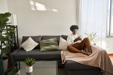Happy relaxed gen z African American teen sitting on couch at home holding cellphone, using mobile...
