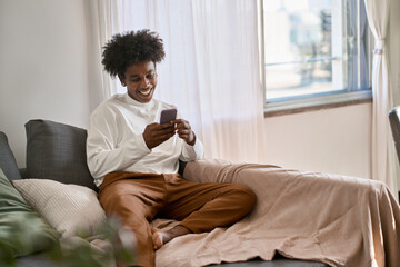 Happy relaxed gen z African American teen sitting on couch at home holding smartphone, using cell...