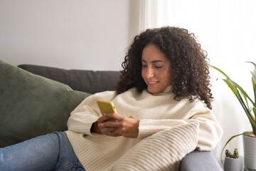 Smiling young latin woman sitting on couch using cell phone at home holding smartphone, looking at...
