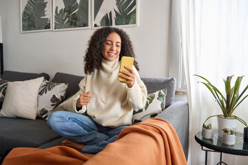 Smiling latin girl sitting on couch using cell phone in living room at home. Relaxed happy young...