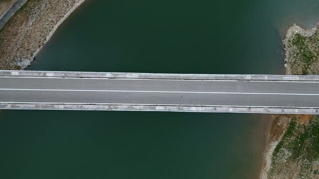 Aerial view of a bridge over a river, the road horizontally separates the image in half, a grey car crosses the frame from left to right, the drone moves down zooming in slightly.