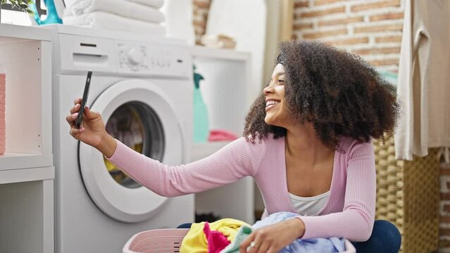 African american woman make selfie by smartphone waiting for washing machine at laundry room