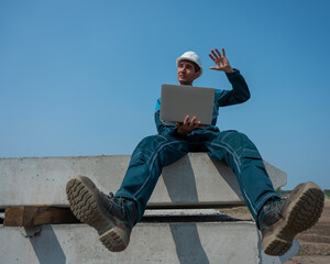 Caucasian male builder in hardhat sits on floor slabs and uses laptop at construction site. 