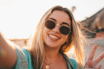 Fototapeta premium Portrait close up of one young beautiful attractive blonde young girl holding camera and taking selfie picture at the beach enjoying outdoors free time with sunset in the background.
