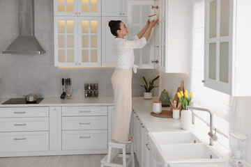 Woman on ladder putting white jar into cupboard at home - Powered by Adobe