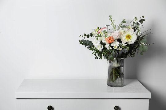 Bouquet with beautiful flowers in vase on chest of drawers near white wall. Space for text