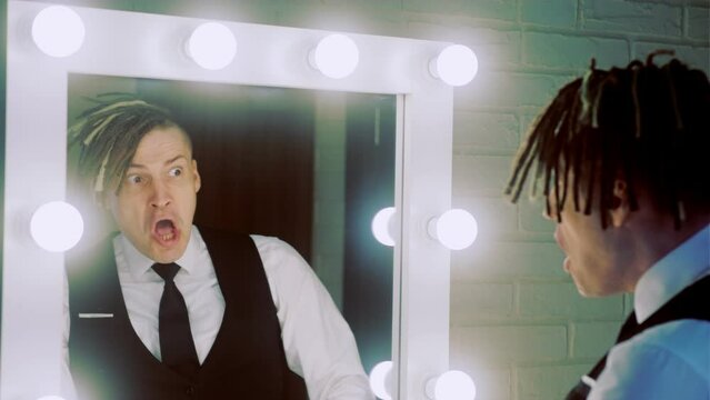 Man with dreadlocks in business suit grimacing, looking in mirror with light bulbs.
