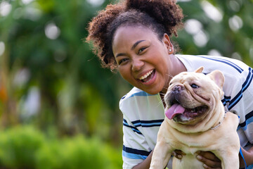 African American woman is playing with her french bulldog puppy while walking in the dog park at grass lawn after having morning exercise during summer