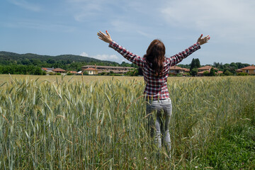 A young woman stands in a wheat field in summer with her back with her arms spread to the sides.