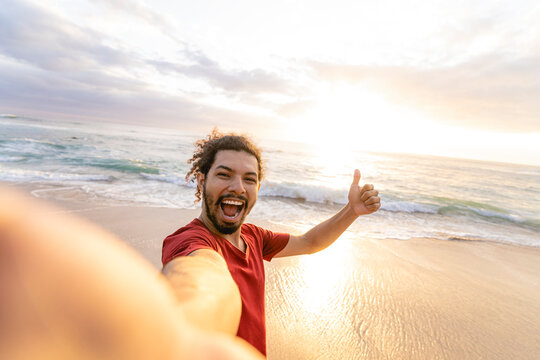 Horizontal image of a very smiling young curly-haired man taking a selfie on the beach with beautiful sunset light behind him. 