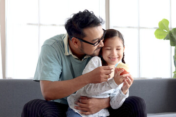 Cute little girl sitting on sofa in living room with her daddy, playing and eating apple together, lovely family, dad and daughter spending time together at home, take care of kid, Happy Father Day.