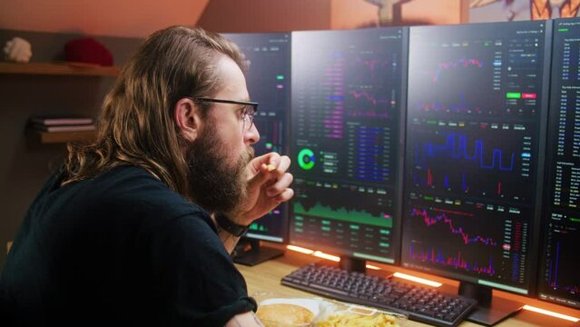 Focused male trader analyzes real-time stocks, exchange market charts on multi-monitor computer workstation, eats fast food. Man works remotely in investment at home office. Cryptocurrency trading.