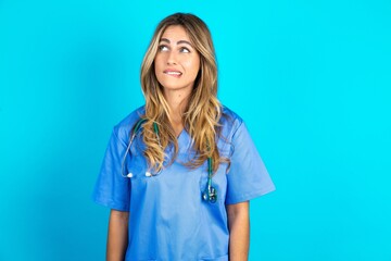 Amazed Young caucasian blonde doctor woman wearing blue uniform bitting lip and looking tricky to...