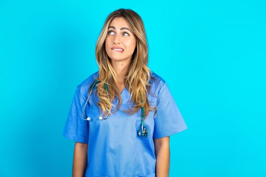 Amazed Young caucasian blonde doctor woman wearing blue uniform bitting lip and looking tricky to empty space.