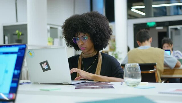 Young black businesswoman sitting at workplace in open space office and working on laptop. Medium handheld shot
