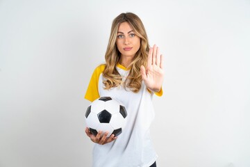Young beautiful woman wearing football T-shirt over white background shows stop sign prohibition symbol keeps palm forward to camera with strict expression