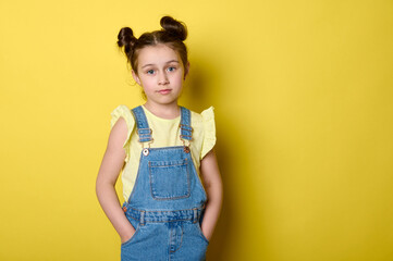 Beautiful smart clever charming little child girl, adorable school kid, pupil in yellow t-shirt and jeans overalls, holding hands in pockets, confidently looking at camera, isolated yellow background