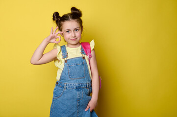 Happy primary school student girl, first grader in blue denim dress overalls and yellow t-shirt, carrying pink backpack, gestures with OK hand sign, cutely smiles looking at camera, yellow background