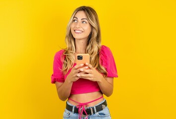 young blonde woman wearing pink crop top over yellow studio background holding in hands mobile phone and looking aside with dreamy look.