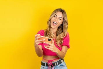 Portrait of an excited young blonde woman wearing pink crop top over yellow studio background playing games on mobile phone.
