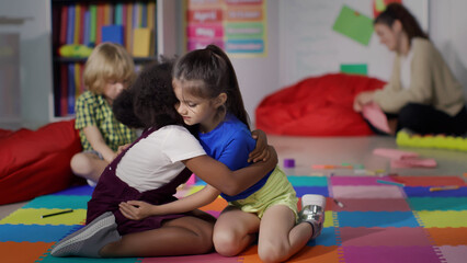 Cute diverse little girls hug sitting on carpet in kindergarten