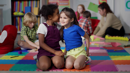 Multiracial little girls tell secrets sitting on floor in kindergarten