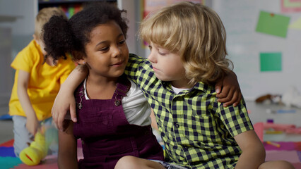 Adorable diverse kids talk and hug sitting on floor in kindergarten playroom