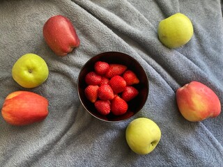 On a gray bedspread, stands a black plate full of ripe and delicious strawberries. Next to it lie ripe apples. The strawberries are red, and the apples are red and yellow.