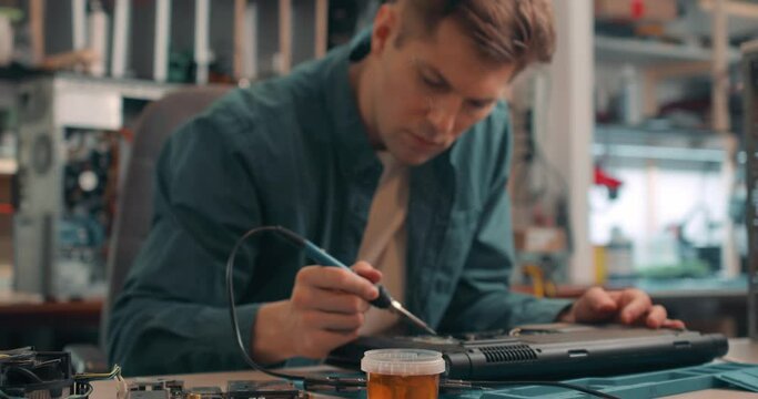 young man working with broken disassembled laptop. Electronic components. Computer analysis. Slow motion attractive man using a soldering iron to fix broken hardware at the repair shop.