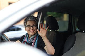 Happy mature woman owner of a new car waving from the driver's seat.