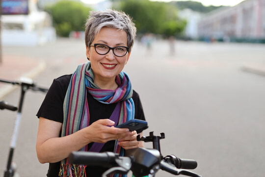 Cheerful mature woman with a smartphone in her hands rents an electric scooter on a city street.