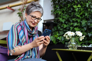 A positive mature woman in glasses with a smartphone in her hands sits at a cafe table.